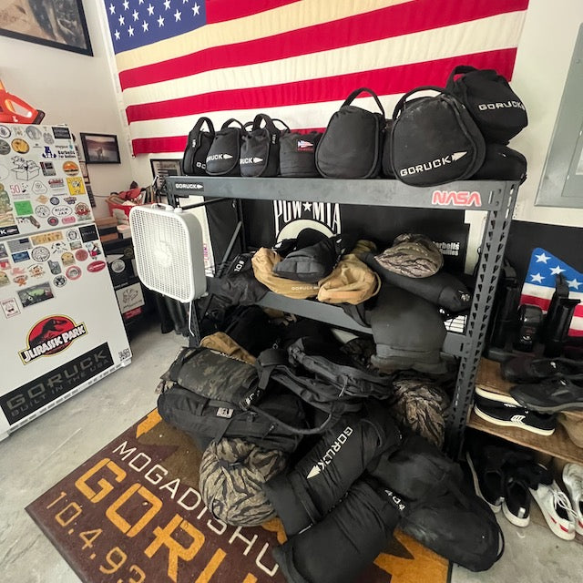 A shelf with GORUCK bags and gear, featuring Sand Kettlebells with padded handles, is set before a large American flag and a sticker-covered fridge.