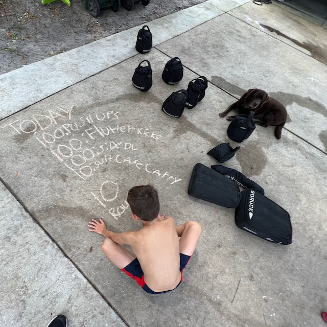 A boy sits on the pavement next to Sand Kettlebells with padded handles, a dog, and a workout plan written in chalk on the ground.