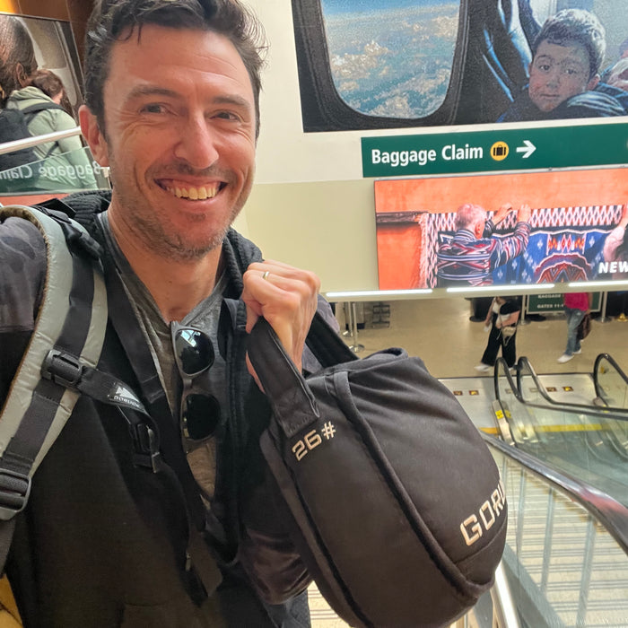 A man smiles on an airport escalator, carrying Sand Kettlebells with a padded handle, his bag over one shoulder, and a baggage claim sign in the background.