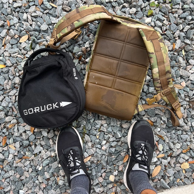Person standing on gravel by a GORUCK backpack, a Sand Kettlebell with padded handle, and a camouflage padded strap, with scattered leaves nearby.