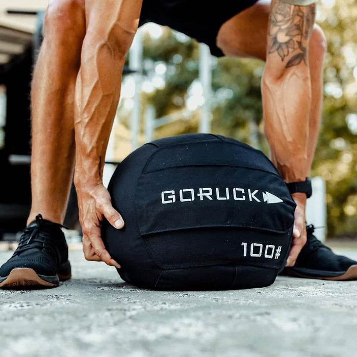 Athlete lifting GORUCK 100 pound sandbag outdoors, rucking gear for fitness training
