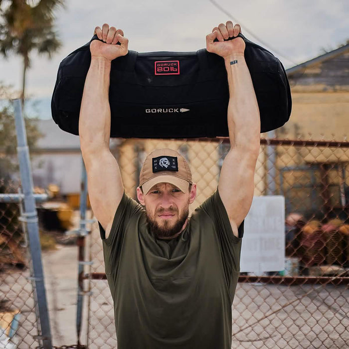 Man lifting GORUCK sandbag overhead outdoors, showcasing tough rucking fitness gear