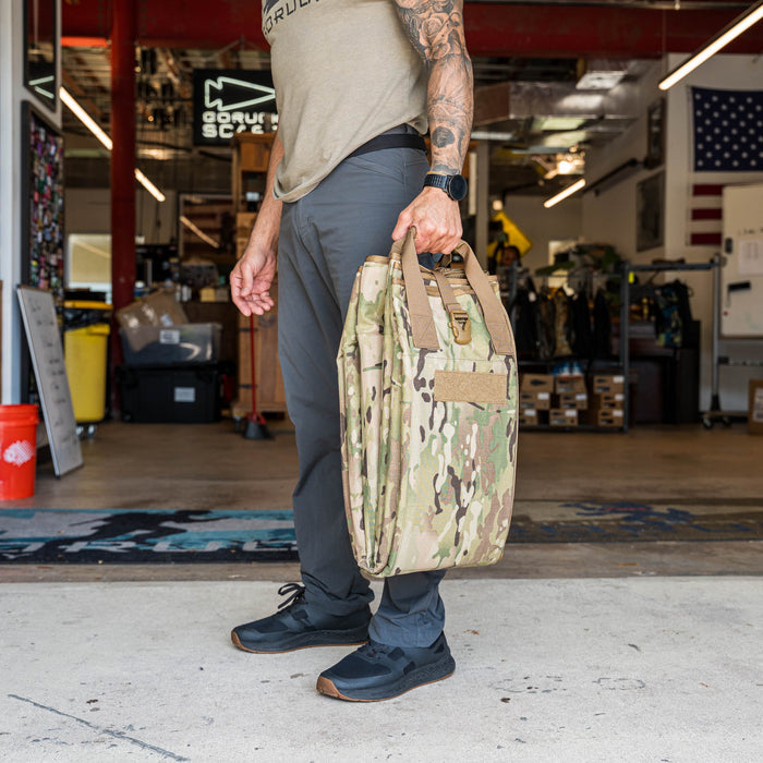 Man holding a camouflage Shooting Mat, standing in a gear shop with boxes, an American flag, and a tripod bracing strap for long range precision visible in the background.