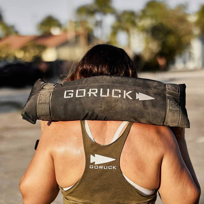 Woman carrying GORUCK sandbag outdoors, wearing GORUCK tank top, fitness training gear
