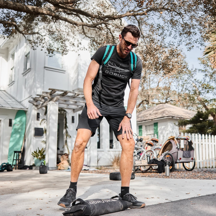 A man in athletic wear stands outside on a driveway, stepping onto the Simple Training Sandbags for a workout, with bikes and trees visible in the background.