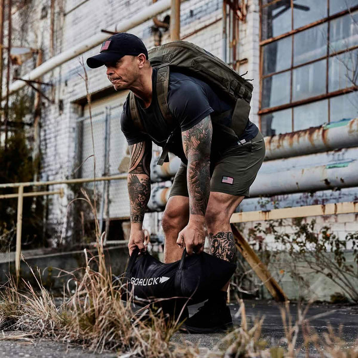 Athlete lifting GORUCK sandbag outdoors, wearing rucksack and workout gear