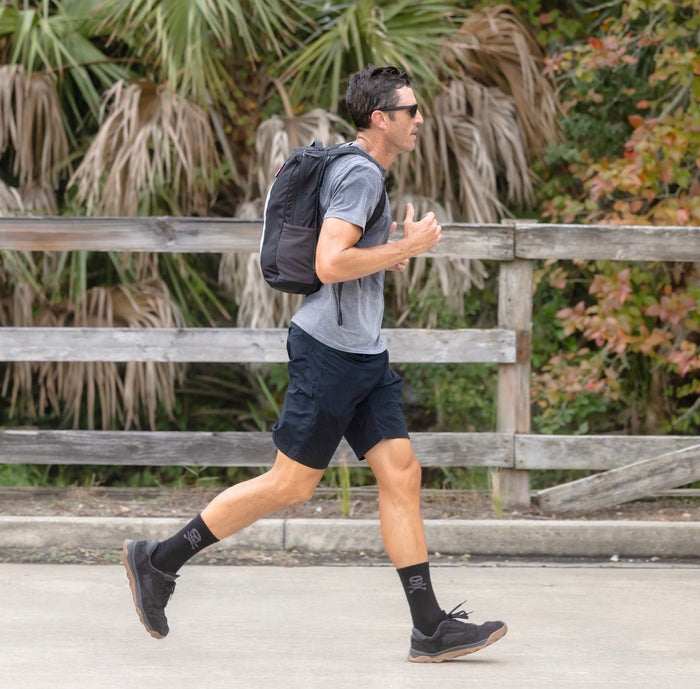 A man jogs on a sidewalk wearing a gray shirt, black shorts, black shoes, and carrying the Basic Rucker backpack.