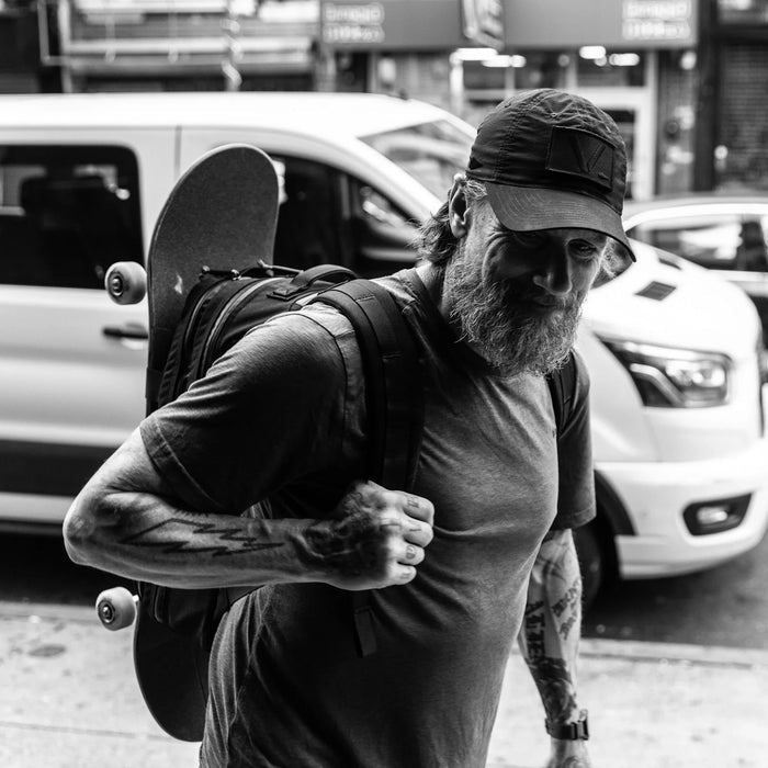 Man with beard and tattoos carrying GORUCK backpack and skateboard on city street