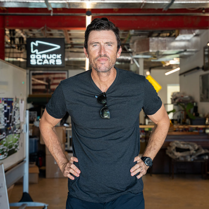 A man wearing the Slick Short Sleeve Tee - Tri-BlendX stands confidently indoors, sunglasses hanging from his shirt and hands on hips. This premium tee provides exceptional comfort and style for any occasion.
