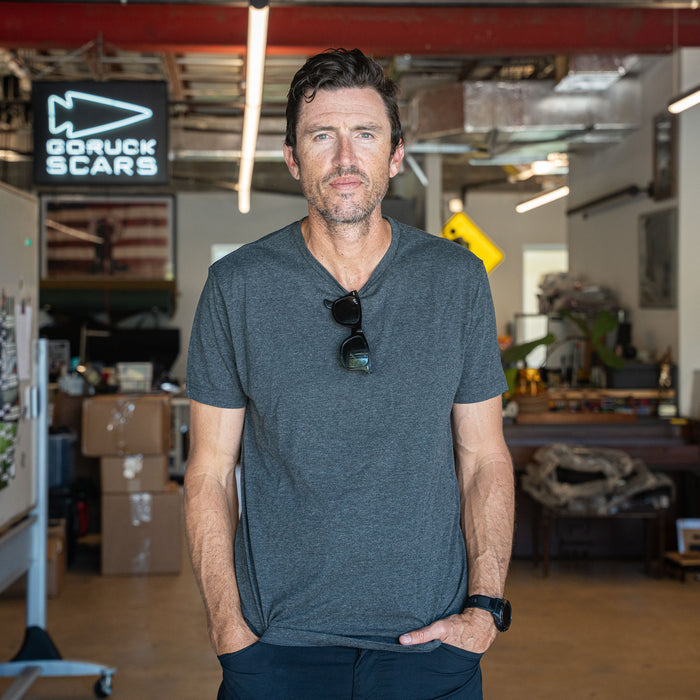A man wearing the Slick Short Sleeve Tee - Tri-BlendX in gray stands in an industrial office, with boxes and a GORUCK SCARS sign behind him.