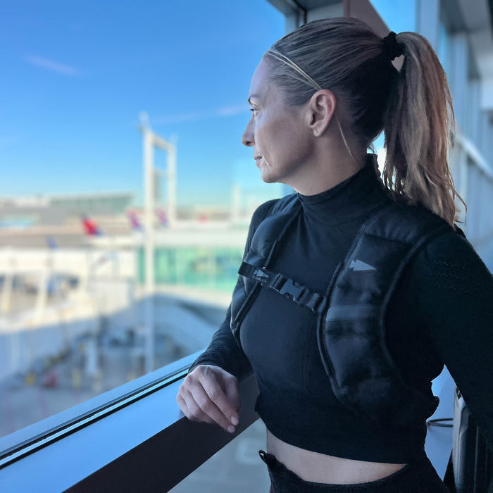 A woman with a ponytail, wearing a black top and the Spy Ruck | Women's Weighted Vest, looks out an airport window with planes visible outside.
