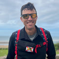 A smiling man by the coast wears sunglasses, a black jacket, and a red star-patterned backpack equipped with the Sternum Pouch - Cordura.