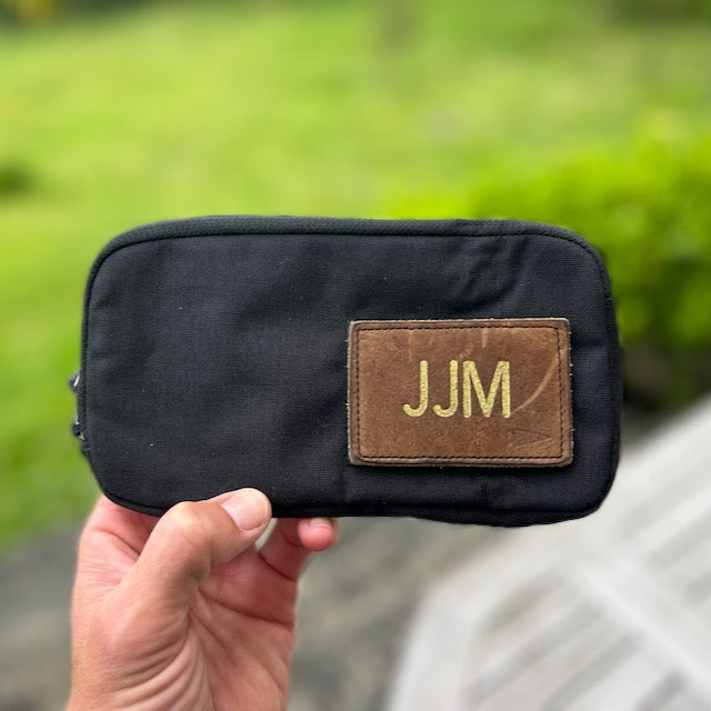 A hand holds the black Sternum Pouch - Cordura with a brown "JJM" patch outdoors against a lush green background.