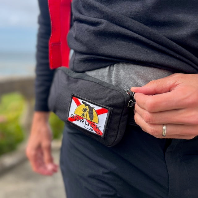A person zips a black Sternum Pouch - Cordura featuring a colorful patch while wearing a wedding ring and casual clothes outdoors.