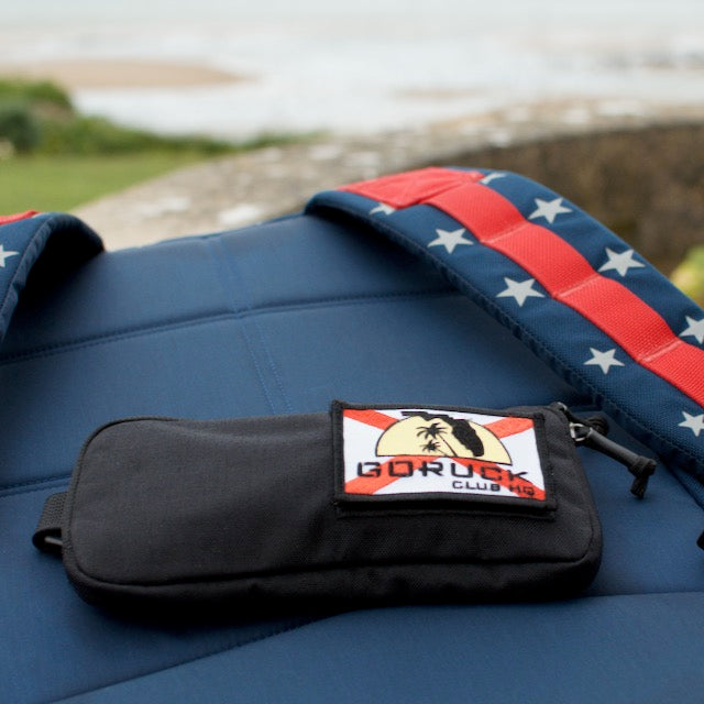 A black Sternum Pouch - Cordura with a GORUCK patch sits on a blue pad with star-patterned red straps outdoors.