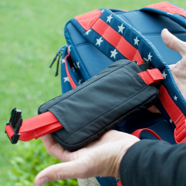 A person attaches a Sternum Pouch - Cordura, in red and black, to a blue backpack with red and white stars.