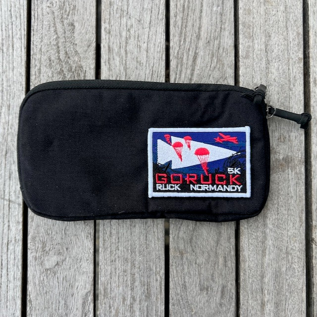 A Sternum Pouch - Cordura in black, shown on wood with a "Goruck Ruck Normandy 5K" patch that features parachutists and a red plane.