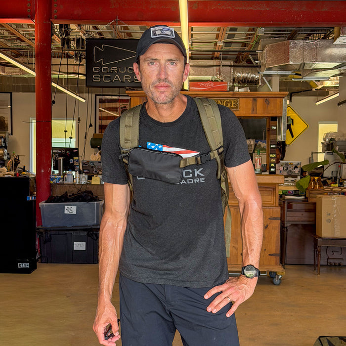 A man indoors wears a black t-shirt and cap, carrying a backpack with the Sternum / Belt Pouch - Waterproof X-PAC featuring an American flag patch for easy access storage.