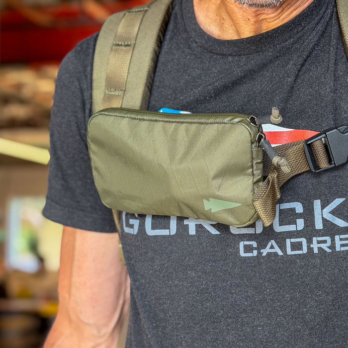 A person wears a black t-shirt and carries the Sternum / Belt Pouch - Waterproof X-PAC in green, attached to their backpack straps for hands-free storage.