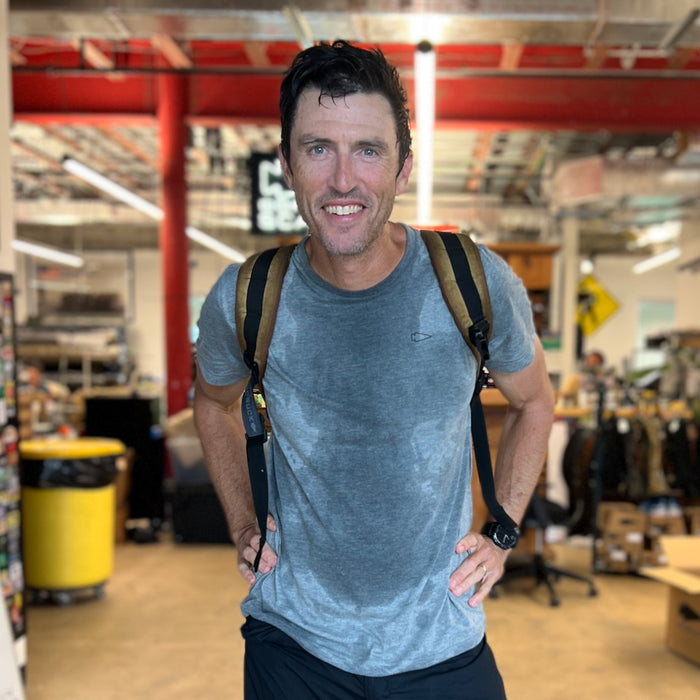 Man smiling in a warehouse, wearing a grey Subtle Spearhead Tee - Unisex Tri-Blend with sweat stains and a backpack, hands on hips.