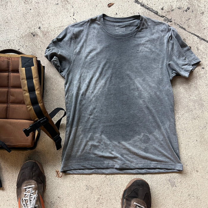 A sweat-stained gray Subtle Spearhead Tee - Unisex Tri-Blend lies on concrete beside a brown backpack and two sneakers.