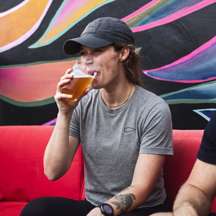 Wearing a Subtle Spearhead Tee - Unisex Tri-Blend and a cap, the person drinks a pint of beer on a red couch in front of a colorful mural.