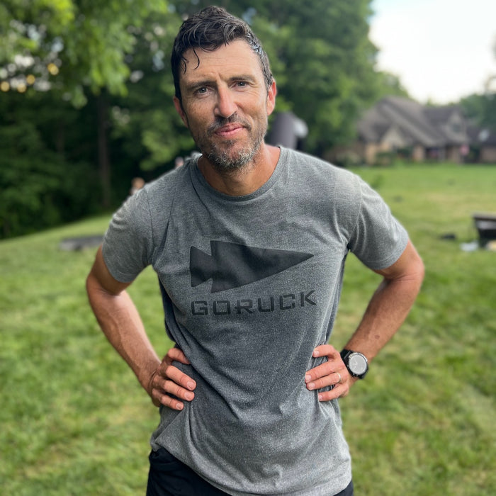 Man in a gray Three Rules Tee - Unisex Tri-BlendX stands outdoors on grass, looking at the camera with hands on hips, ready to look cool and never get lost.