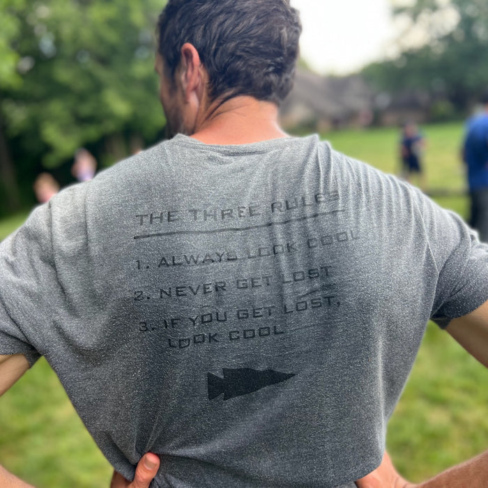 A man stands outdoors wearing the Three Rules Tee - Unisex Tri-BlendX, featuring three rules on the back like "Always look cool" and "Never get lost.