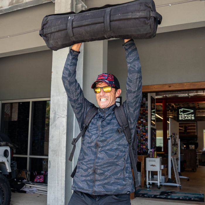 A man in sunglasses lifts a large black duffel bag overhead outside a store, smiling with effort, his Warm Up Full Zip jacket with zippered pockets keeping his essentials secure.