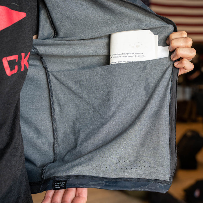 A person shows a gray Warm Up Full Zip jacket with lightweight ToughDry shoulders, featuring a large inside pocket holding a folded piece of paper.