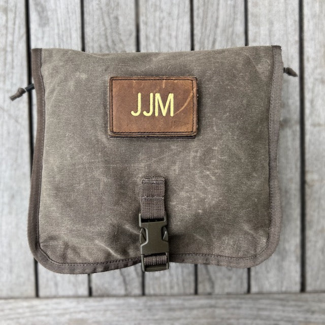 The Wire Dopp - Waxed Canvas pouch, featuring a leather "JJM" patch and buckle, is perfect for storing EDC essentials and is displayed on a wooden background.