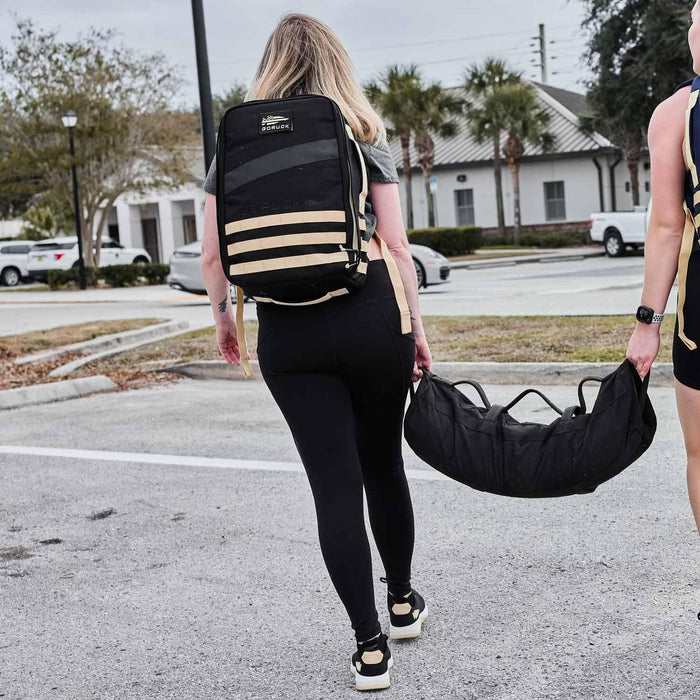 Woman rucking outdoors with GORUCK backpack and sandbag, demonstrating tough rucking gear.