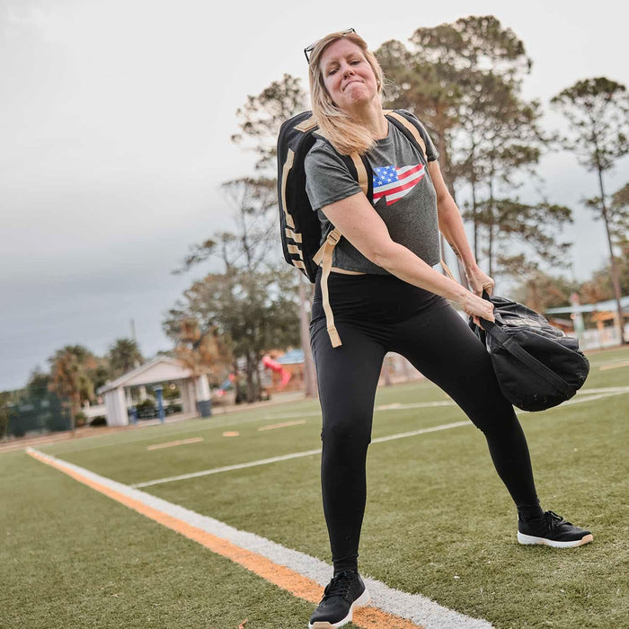 Woman rucking on a sports field wearing GORUCK gear and lifting a sandbag for training