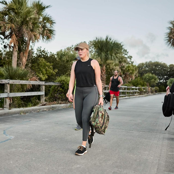 Woman rucking with GORUCK gear on outdoor path, wearing athletic apparel and carrying camo backpack