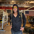 A man stands indoors before an American flag, wearing sunglasses, a backpack, and the Men's USA Sun Hoodie - Full-Zip - ToughMesh.