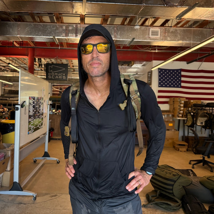 A man stands indoors before an American flag, wearing sunglasses, a backpack, and the Men's USA Sun Hoodie - Full-Zip - ToughMesh.