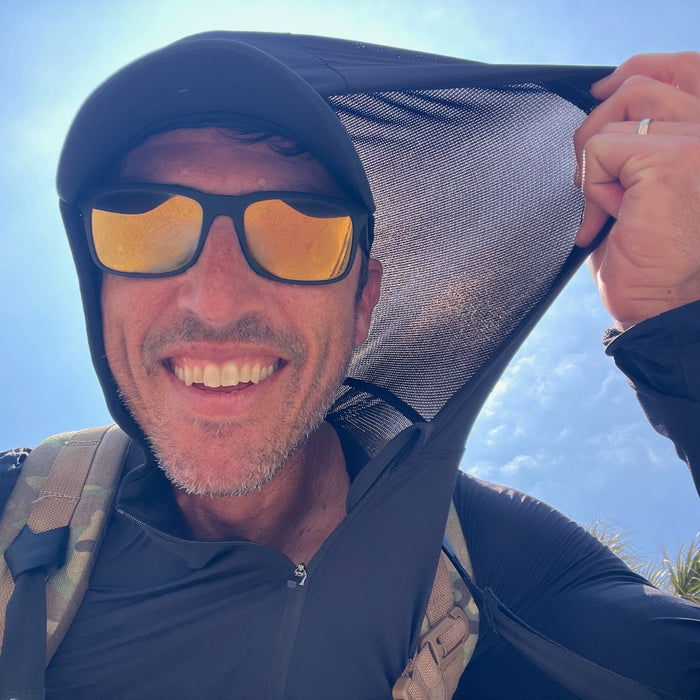 A smiling man in sunglasses lifts the hood of his Men's USA Sun Hoodie - Full-Zip - ToughMesh outdoors on a sunny day, with the sky in the background.