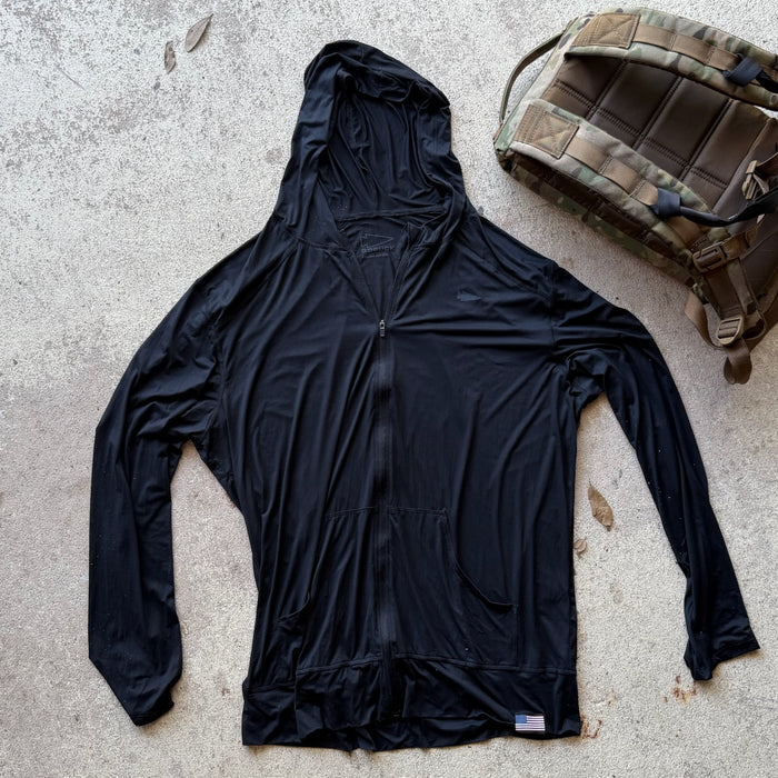 A Men's USA Sun Hoodie - Full-Zip - ToughMesh in black is laid flat beside a camouflage backpack on concrete.
