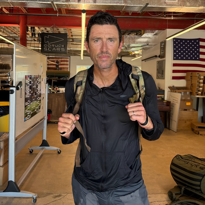 A man wearing the Men's USA Sun Hoodie - Full-Zip - ToughMesh stands indoors with a backpack; an American flag hangs on the wall behind him.