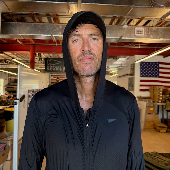 A man wears the Men's USA Sun Hoodie - Full-Zip - ToughMesh indoors, with an American flag and shelves in the background.